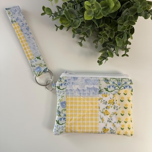 May include: A patchwork coin purse and wristlet in blue, yellow, and white floral and gingham patterns. The purse has a zip closure and a silver metal ring to attach the wristlet. The wristlet strap is patterned to match the purse. A green plant is in the background.