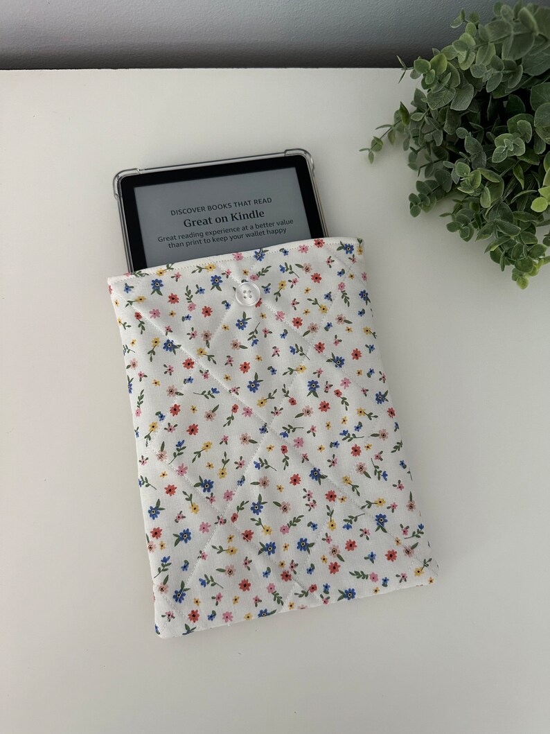 Op de afbeelding: Een witte gewatteerde Kindle-hoes met een bloemenpatroon in rood, blauw en geel. De hoes bevat een Kindle met de tekst "Great on Kindle". De hoes is gesloten met een witte knoop.