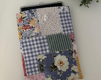 May include: A rectangular fabric sleeve for a tablet featuring a patchwork design of various floral, gingham, and striped patterns in shades of blue, white, pink, and yellow. The sleeve is quilted and secured with a single clear plastic button.