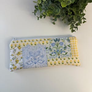 May include: A rectangular, quilted pencil case with a zipper closure. The case features a patchwork design with floral and patterned fabric in yellow, white, and blue. The case is approximately 8 inches long.