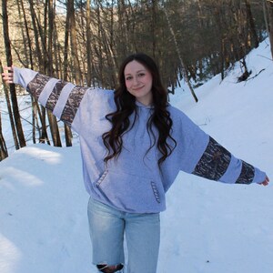 May include: A light blue hoodie with camouflage patterned sleeves. The hoodie has a front pocket and is paired with light wash jeans with distressed knees. The background shows a snowy forest.