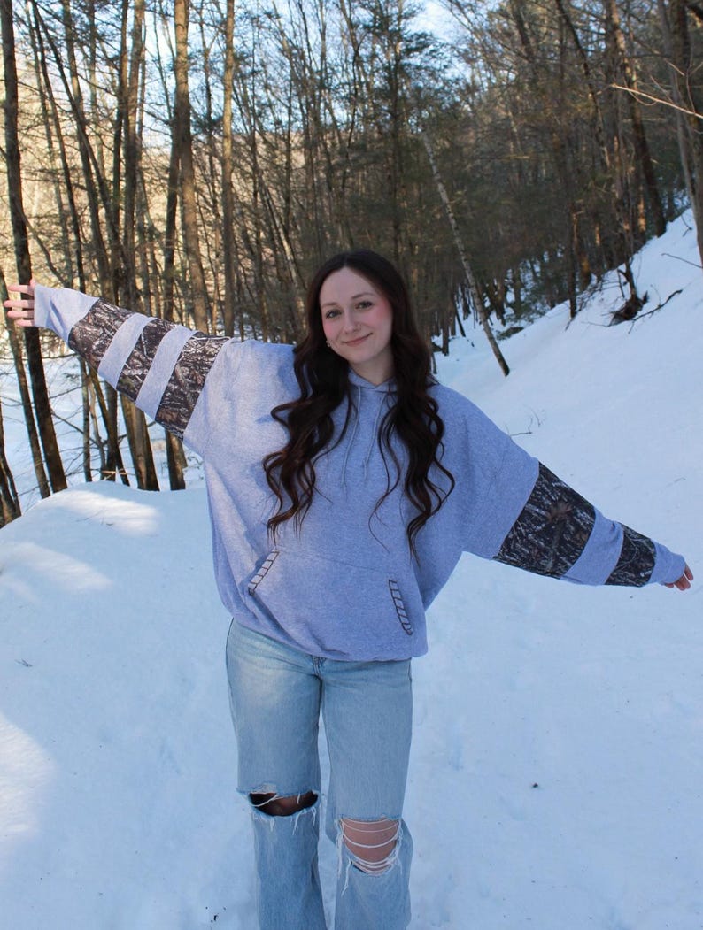 May include: A light blue hoodie with camouflage patterned sleeves. The hoodie has a front pocket and is paired with light wash jeans with distressed knees. The background shows a snowy forest.