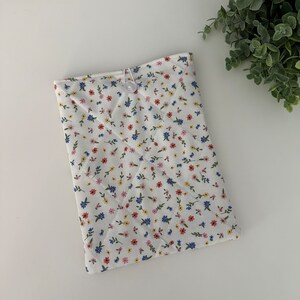 Op de afbeelding: Een witte gewatteerde tablethoes met een bloemenpatroon in rood, blauw en geel. De hoes is vastgezet met een witte knoop en een elastische lus. Het bloemenpatroon is een herhalend ontwerp van kleine bloemen en groene bladeren.