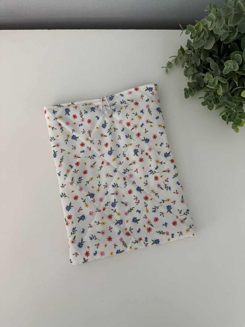 Op de afbeelding: Een witte gewatteerde tablethoes met een bloemenpatroon in rood, blauw en geel. De hoes is vastgezet met een witte knoop en een elastische lus. Het bloemenpatroon is een herhalend ontwerp van kleine bloemen en groene bladeren.