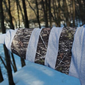 May include: A gray sweatshirt sleeve with vertical camouflage fabric stripes. The camouflage pattern features shades of brown, green, and black. The background shows a snowy landscape and trees.