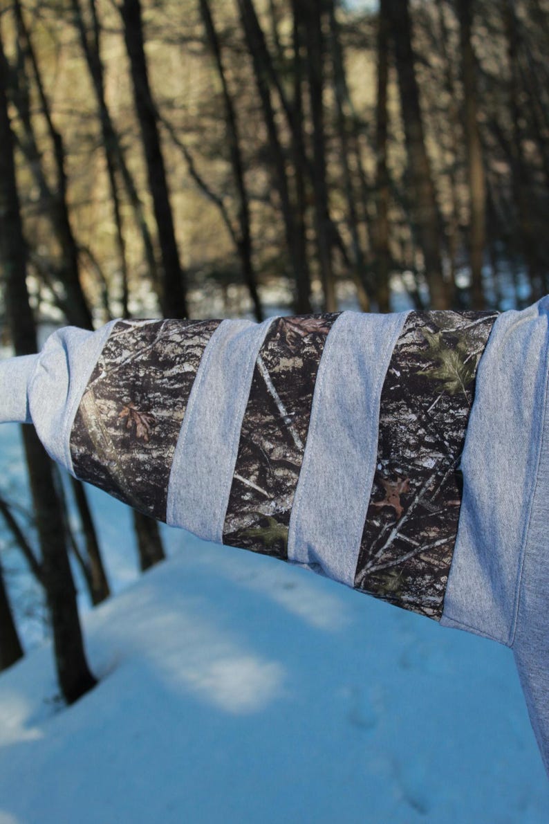 May include: A gray sweatshirt sleeve with vertical camouflage fabric stripes. The camouflage pattern features shades of brown, green, and black. The background shows a snowy landscape and trees.