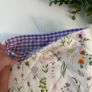 May include: A quilted oven mitt with a floral pattern featuring various wildflowers in shades of purple, orange, and pink on a white background. The interior lining is a purple and white gingham check. A white loop is attached for hanging.