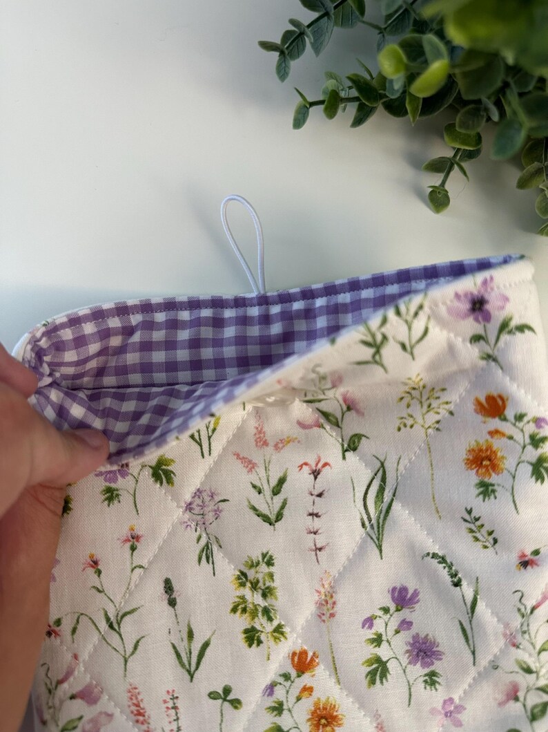 May include: A quilted oven mitt with a floral pattern featuring various wildflowers in shades of purple, orange, and pink on a white background. The interior lining is a purple and white gingham check. A white loop is attached for hanging.