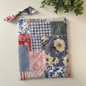May include: A patchwork quilted pouch with a zip closure and a fabric pull. The pouch features floral, gingham, and striped fabric patterns in blue, pink, and red. A matching fabric bow is attached to the zip.