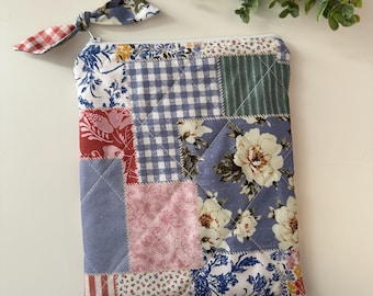 May include: A patchwork quilted pouch with a zipper closure and a fabric pull. The pouch features a variety of floral, gingham, and striped fabric patterns in shades of blue, pink, and red. A matching fabric bow is attached.