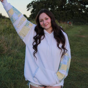 Peut inclure: Sweat à capuche blanc avec des manches patchwork jaunes, bleues et blanches. Le sweat à capuche a une poche avant et une capuche à cordon. Les manches présentent un motif unique.