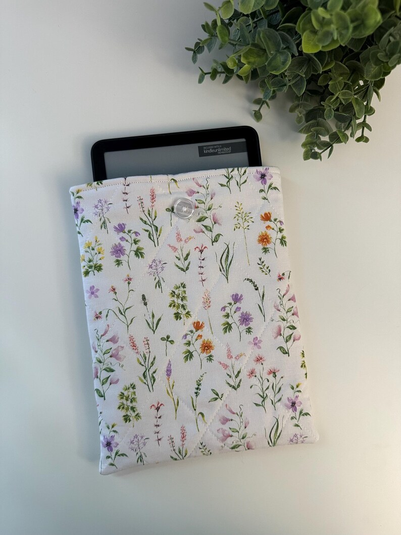May include: White quilted Kindle cover with a floral pattern in shades of purple, orange, and pink. The cover has a button closure and a Kindle device is partially visible inside. The cover is designed to protect e-readers.