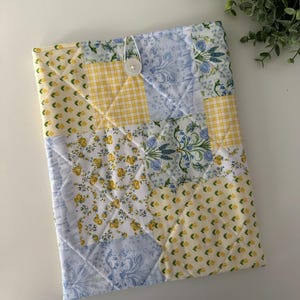 Peut inclure: Une housse de livre en tissu matelassé avec un motif patchwork dans les tons jaunes et bleus. La housse présente une variété de motifs floraux et un seul bouton blanc.