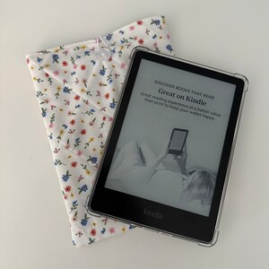 Op de afbeelding: Een Kindle e-reader met een doorzichtige beschermhoes ligt naast een witte stoffen hoes met bloemenprint. De e-reader toont de tekst "Great on Kindle". De hoes is gewatteerd en vastgemaakt met een knoop.