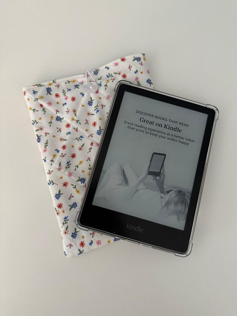 Op de afbeelding: Een Kindle e-reader met een doorzichtige beschermhoes ligt naast een witte stoffen hoes met bloemenprint. De e-reader toont de tekst "Great on Kindle". De hoes is gewatteerd en vastgemaakt met een knoop.