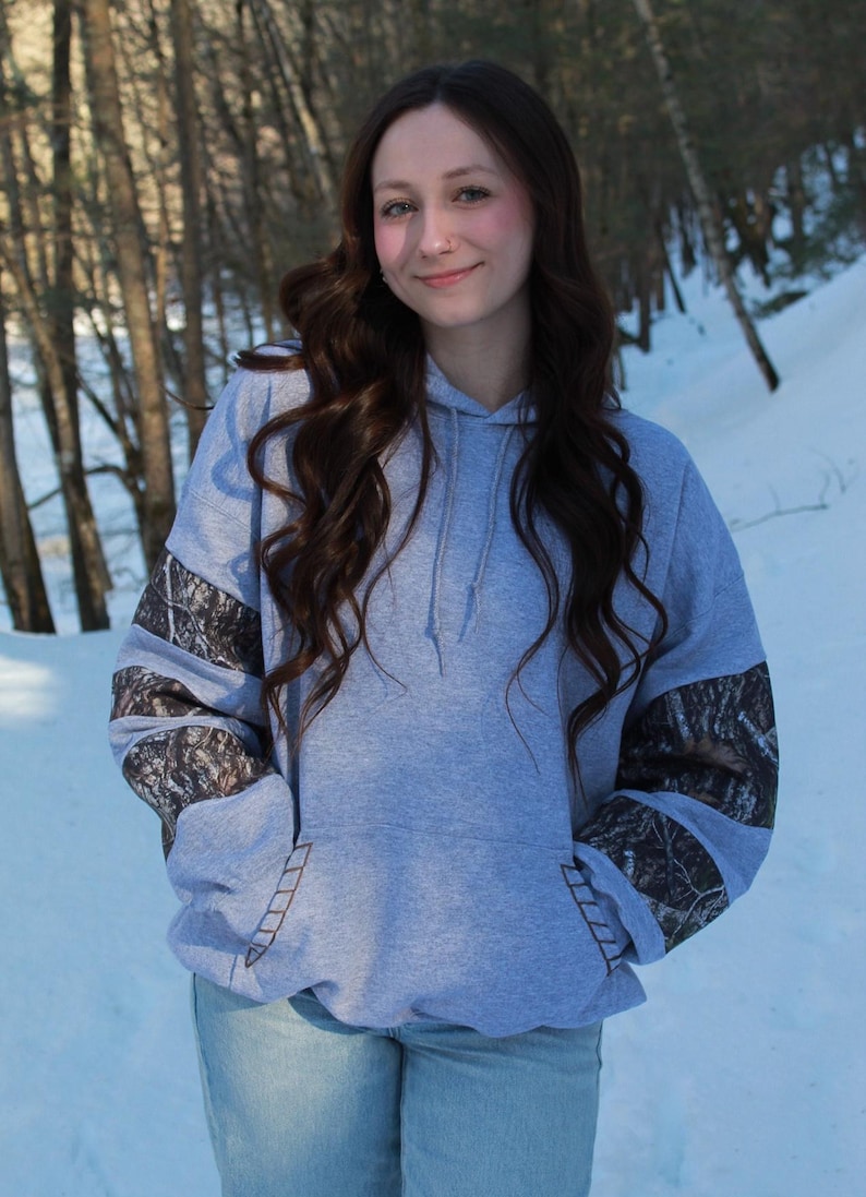 May include: A light gray hoodie with a front pocket and a drawstring hood. The sleeves feature a camouflage pattern. The person is wearing blue jeans. The background is a snowy forest.