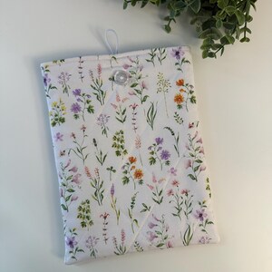 May include: A white quilted tablet sleeve featuring a colorful floral pattern with lavender, orange, and purple flowers. The sleeve has a button closure and a loop for hanging.