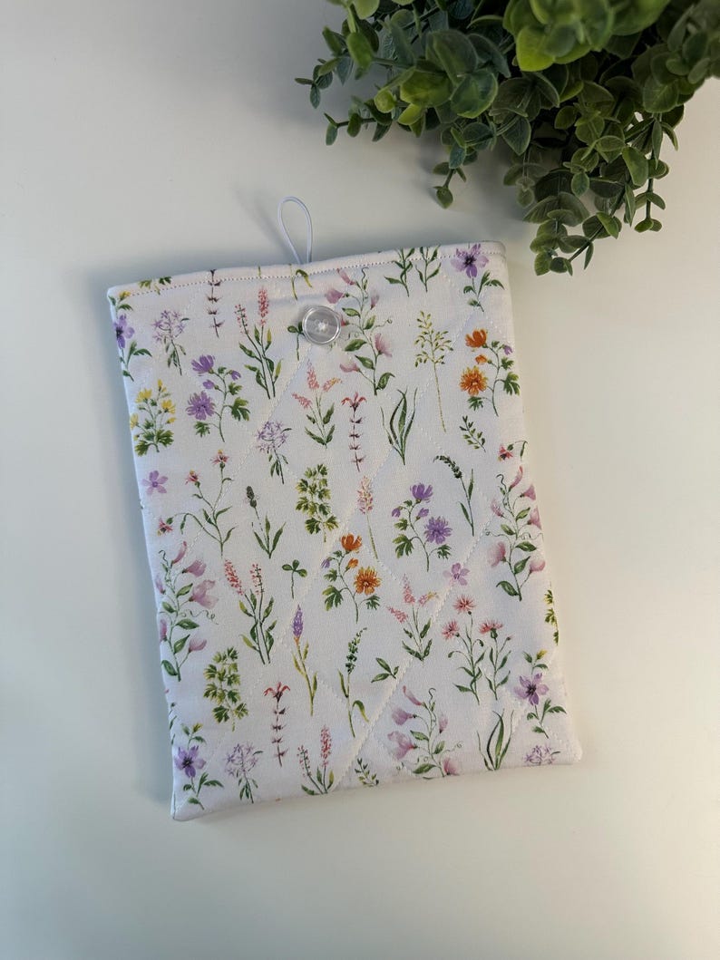 May include: A white quilted tablet sleeve featuring a colorful floral pattern with lavender, orange, and purple flowers. The sleeve has a button closure and a loop for hanging.
