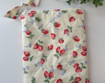 May include: Cream-colored quilted pouch with a zipper closure and a matching bow. The pouch is decorated with a pattern of red strawberries and blue flowers. The bow is attached to the zipper.