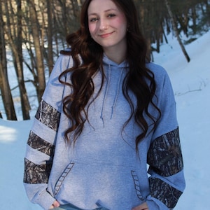 May include: A light gray hoodie with a front pocket and a drawstring hood. The sleeves feature a camouflage pattern. The person in the image is wearing blue jeans and is standing in front of a snowy background with trees.
