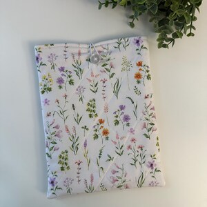 May include: A white quilted tablet sleeve with a floral pattern featuring purple, pink, yellow, and orange wildflowers. The sleeve has a button closure and is designed to protect a tablet. The overall design is a cottagecore aesthetic.