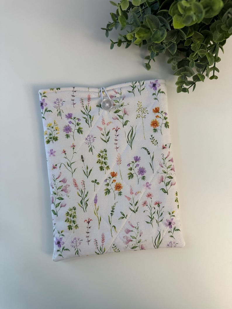 May include: A white quilted tablet sleeve with a floral pattern featuring purple, pink, yellow, and orange wildflowers. The sleeve has a button closure and is designed to protect a tablet. The overall design is a cottagecore aesthetic.