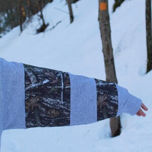 May include: A gray sweatshirt sleeve with alternating panels of solid gray and camouflage fabric. The camouflage pattern features shades of brown, green, and black. The background shows a snowy forest scene.