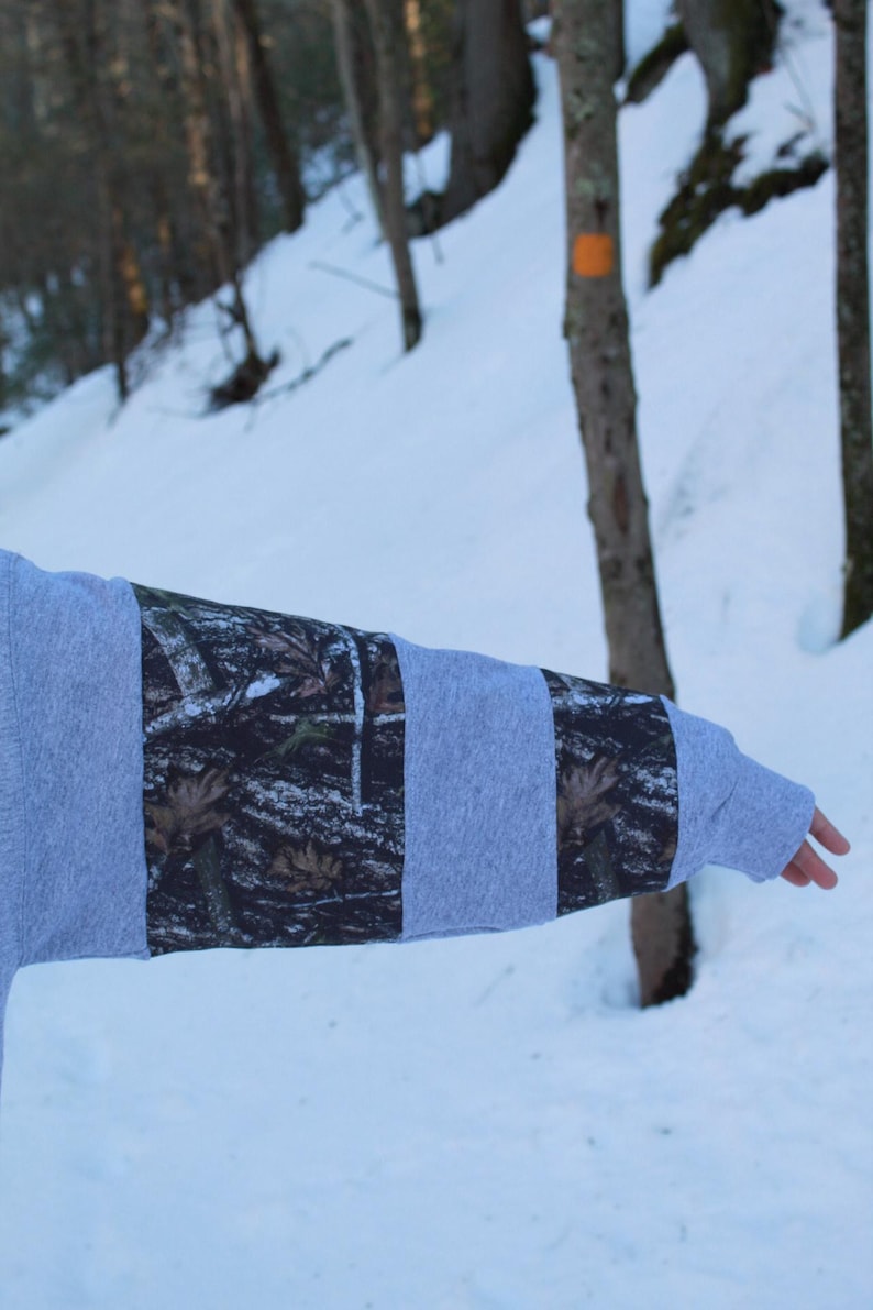 May include: A gray sweatshirt sleeve with alternating panels of solid gray and camouflage fabric. The camouflage pattern features shades of brown, green, and black. The background shows a snowy forest scene.