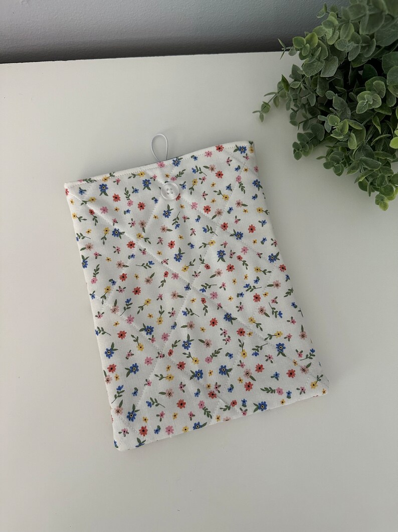 Op de afbeelding: Een witte gewatteerde tablethoes met een bloemenpatroon in rood, blauw en geel. De hoes heeft een knoopsluiting en een lus aan de bovenkant. Het bloemenpatroon staat tegen een witte achtergrond.