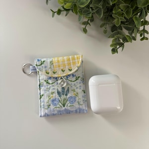 May include: A small, quilted pouch with a floral pattern in blue and white, and a yellow gingham flap. It has a silver key ring attached and a white button closure. Next to it is a white, rectangular case.