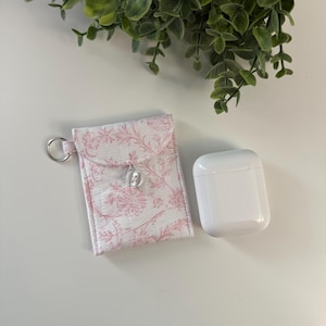 May include: A white and pink floral print fabric case with a silver key ring next to a white wireless earbud charging case. The fabric case has a button closure. Green foliage is in the background.