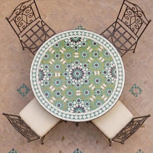 Könnte beinhalten: Draufsicht auf einen runden Gartentisch und Stühle. Der Tisch hat eine Mosaikplatte mit grünen, blauen und weißen geometrischen Mustern. Vier schmiedeeiserne Stühle mit cremefarbenen Kissen umgeben den Tisch. Ein passender Beistelltisch ist ebenfalls zu sehen.