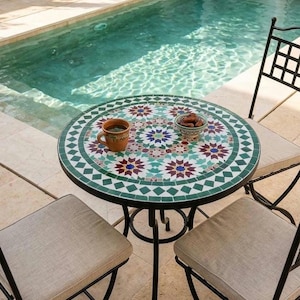 May include: A round mosaic tile table with a black metal frame and three matching chairs. The table has a green, white, red, and blue floral pattern. A cup of coffee and a bowl of dates sit on the table near a swimming pool.