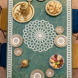 May include: An overhead shot of a green mosaic table set for tea. Gold teapots, plates, and small bowls are arranged on the table, alongside pastries. The table features a white geometric pattern in the centre.