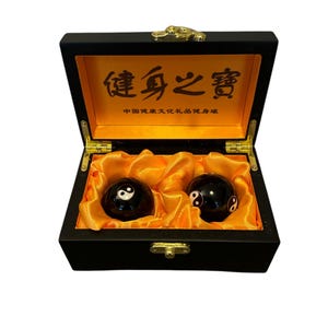 Op de afbeelding: Twee zwarte yin yang ballen in een zwarte houten doos met een gouden sluiting. De doos is gevoerd met oranje satijn en het deksel heeft de Chinese karakters "健康之寶" die vertaald worden als "Schat van gezondheid".