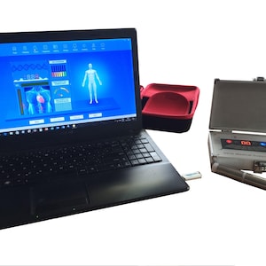 Puede incluir: Un portátil negro que muestra una interfaz de escaneo corporal médico con una figura humana. También hay un dispositivo médico plateado con una pantalla digital y un estuche de transporte rojo y negro.