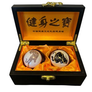Può includere: Una scatola di legno nero con una fibbia dorata e un'iscrizione dorata in cinese che recita "健身之寶" (Tesoro della salute). La scatola è aperta e contiene due sfere di ceramica bianca con disegni di animali dipinti. Una sfera ha un disegno di tigre e l'altra ha un disegno di tartaruga.