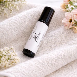 May include: A black essential oil roller bottle with a white label reading "Relax" in cursive. The bottle rests on a white towel, with small white and pink flowers in the background. The product is from Twinkle Wellness.
