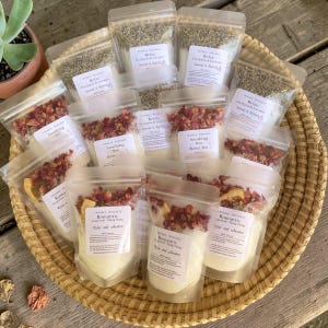 May include: A collection of clear, resealable pouches filled with bath salts, dried flower petals, and botanical elements. Each pouch features a white label with the brand name Twinkle Wellness, product names like Relax, Soothing, and Romantic, and ingredients.