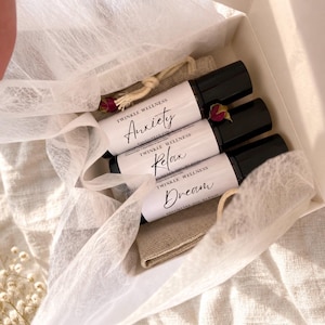 May include: Three glass roller bottles with black caps and white labels in a gift box. The labels read Twinkle Wellness Anxiety, Relax, and Dream. Each bottle contains an essential oil blend and is nestled in a beige fabric pouch with dried rosebuds.