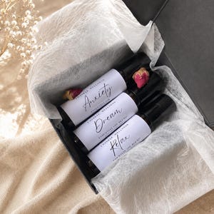 May include: Three dark glass roller bottles with white labels in a black gift box lined with white tissue paper. The labels read Twinkle Wellness Anxiety, Dream, and Relax. Small dried pink rosebuds are tucked between the bottles in the box.