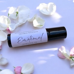 May include: A dark amber glass roller bottle with a white label featuring the text Twinkle Wellness, Emotional, and Essential Oil. The bottle is surrounded by scattered white flower petals with subtle pink accents on a light purple surface.