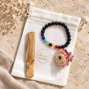May include: A white drawstring pouch with the text "twinkle WELLNESS" printed in gold, containing a black beaded bracelet with multi-coloured chakra stones, a single piece of light brown palo santo wood, and a dried pink and white flower.