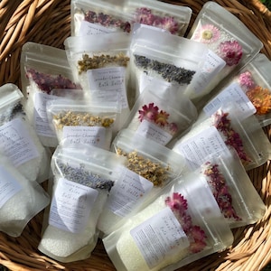 May include: A collection of clear, resealable pouches filled with white bath salts and topped with dried flowers and herbs. Each pouch features a white label with the Twinkle Wellness brand name and specific scent names like Lavender-Chamomile, Relax, and Sweet Dream.
