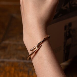 May include: A gold-toned metal bracelet with a nail-shaped design. The bracelet is worn on a person's wrist.