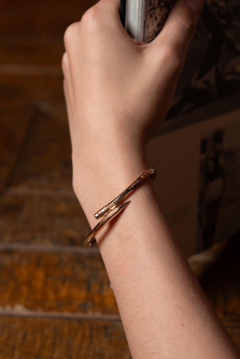 May include: A gold-toned metal bracelet with a nail-shaped design. The bracelet is worn on a person's wrist.