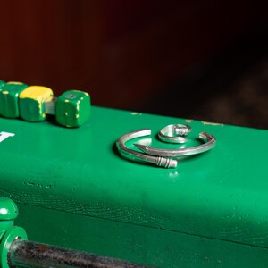May include: Two silver rings, one a simple band and the other a nail-shaped design, are displayed on a green surface.