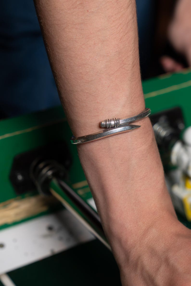May include: A silver bracelet with a unique design featuring a curved band and a small, textured detail.