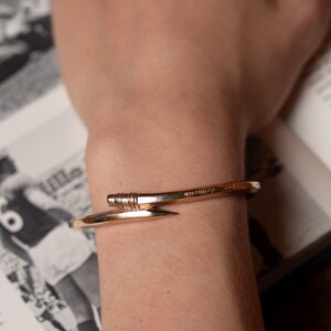 May include: A gold-toned metal bracelet with a nail-shaped design. The bracelet is engraved with the word "nowher" on the top.