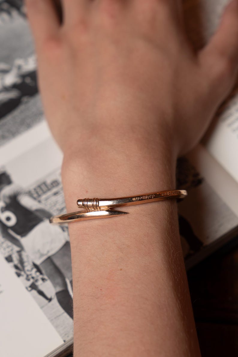 May include: A gold-toned metal bracelet with a nail-shaped design. The bracelet is engraved with the word "nowher" on the top.